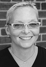 Black and white photo of Jan Beatty wearing rectangular light colored glasses. Her hair is cropped short. She is smiling pleasantly and wears small silver earrings, a chain necklace, and a black v-neck shirt.