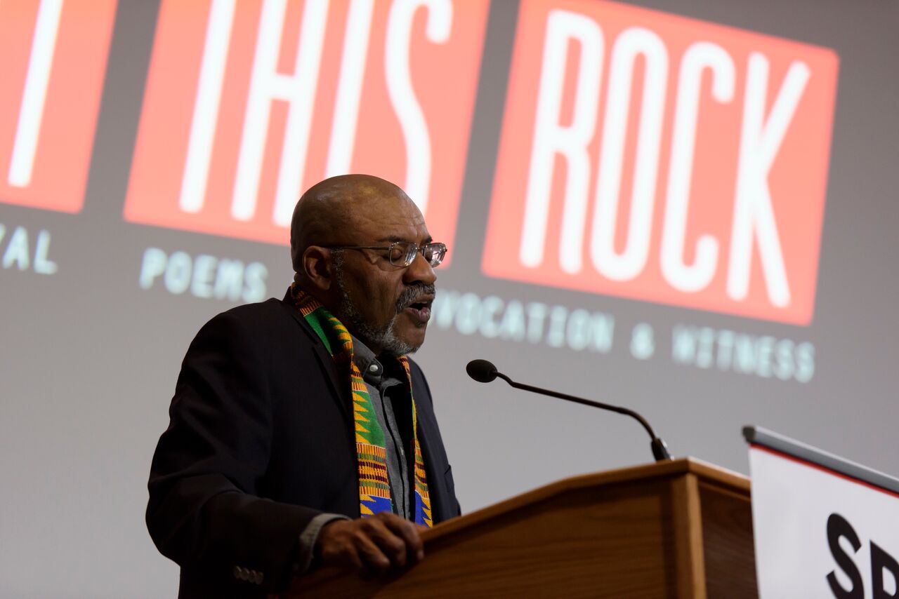 Image of Kwame Dawes at the microphone during his reading at Split This Rock Poetry Festival 2018. He wears a dark jacket, a kinte clothe scarf, and glasses.