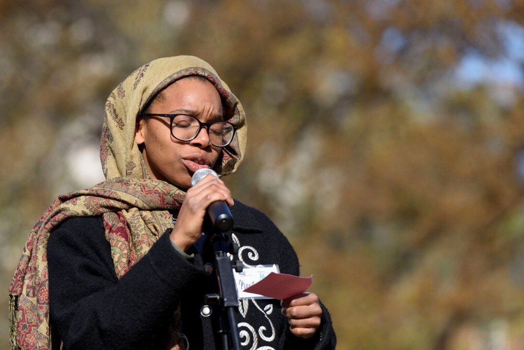 Image of a person sharing a poem at the 2018 festival public action. The person has brown skin and glasses, wears a head wrap, and holds a microphone.
