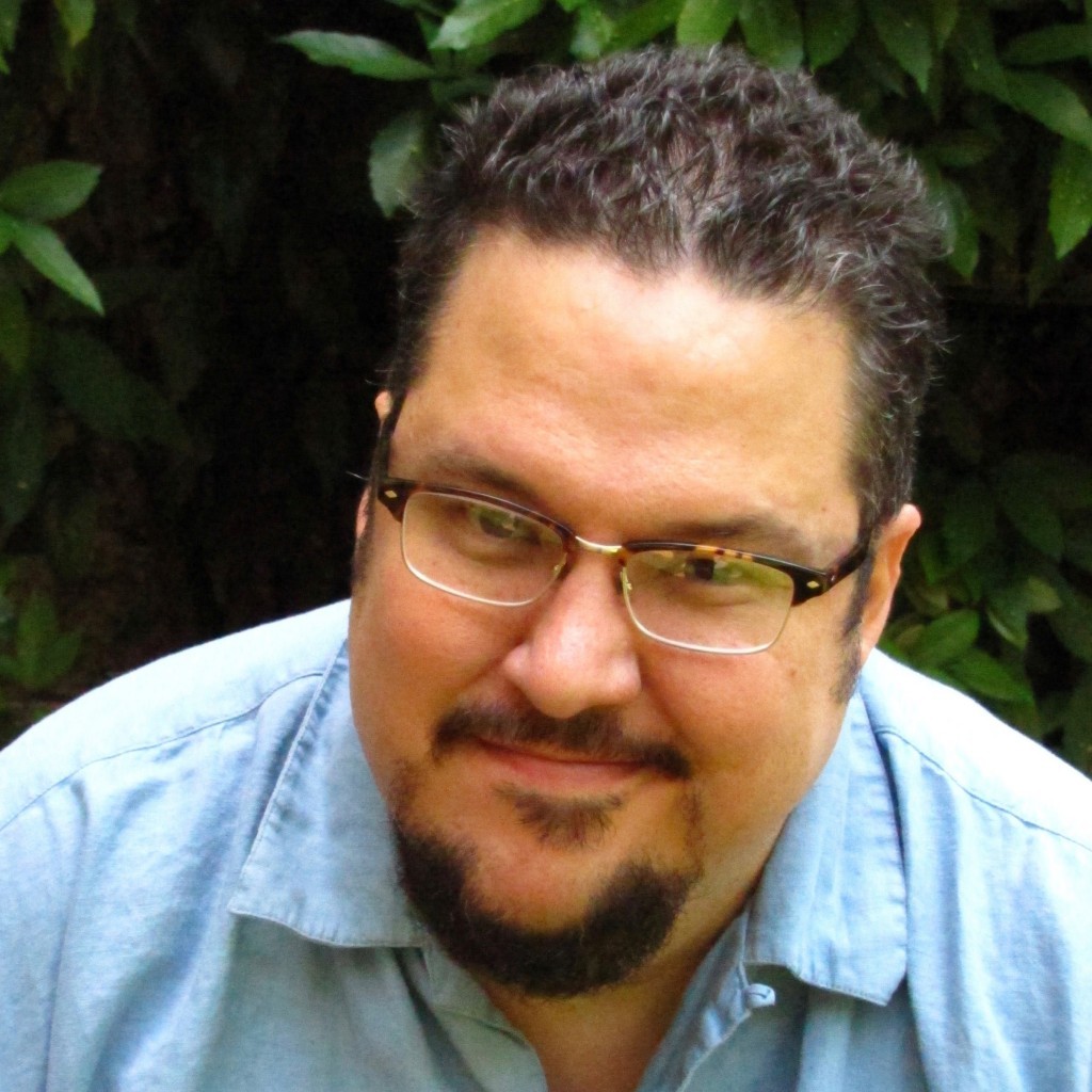 A latino man with brown hair and glasses leans slightly into the camera with a smile on his face and wearing a light blue shirt