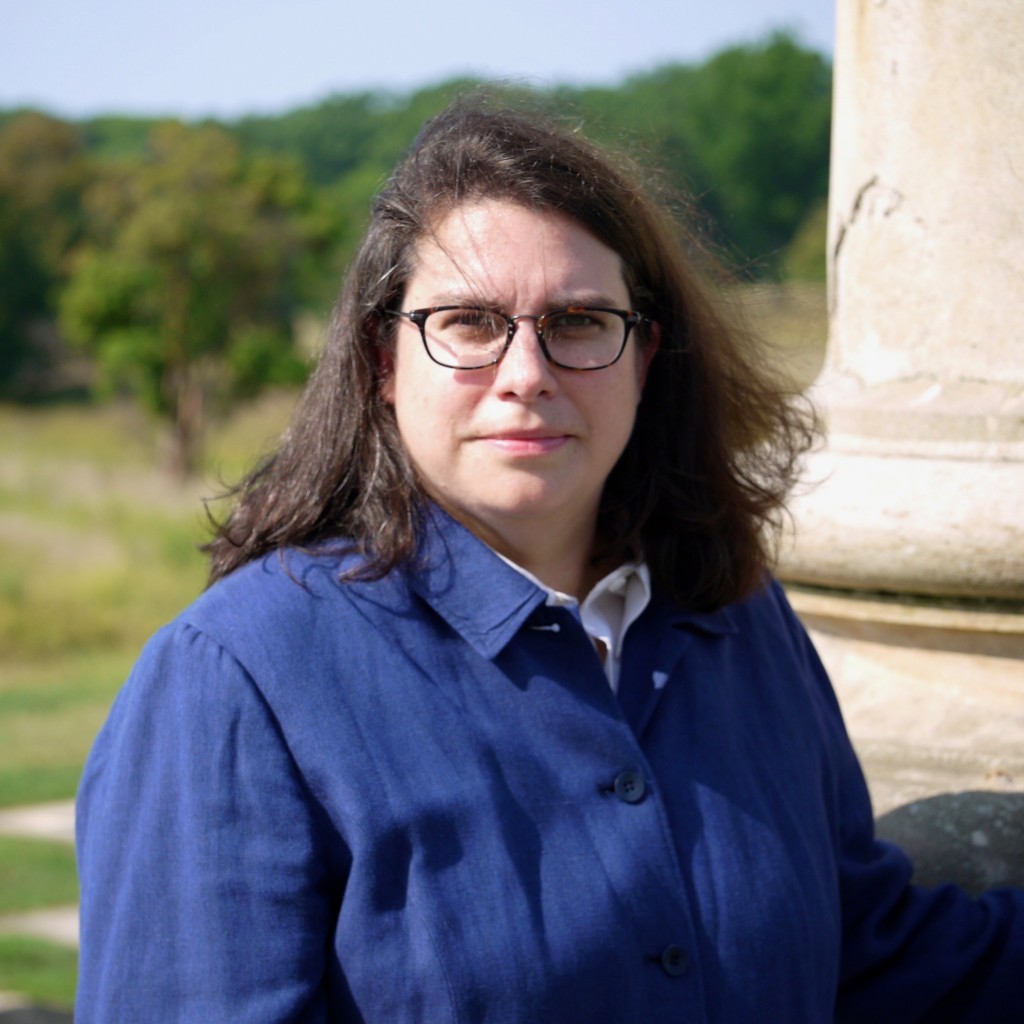 A white woman with brown hair and glasses wearing a blue shirt passively smiles into the camera while standing in a nature area