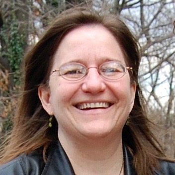 A white woman with brown hair and glasses smiles broadly with trees in the background