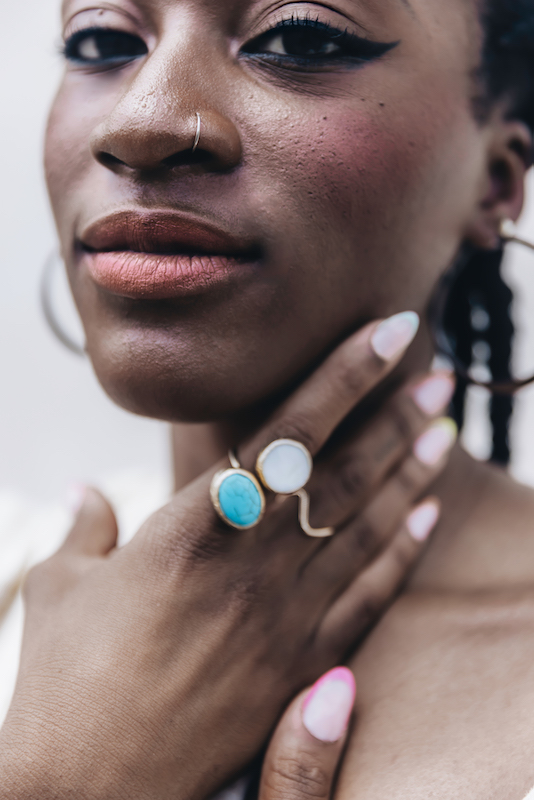 Alexa is Black and has black cornrows. She stares directly at the camera with a closed mouth, half smile. She has a hoop nose ring, and winged eyeliner. Her hand has brightly-colored nails, a turquoise ring and a gold ring, and rests lightly on her chin.