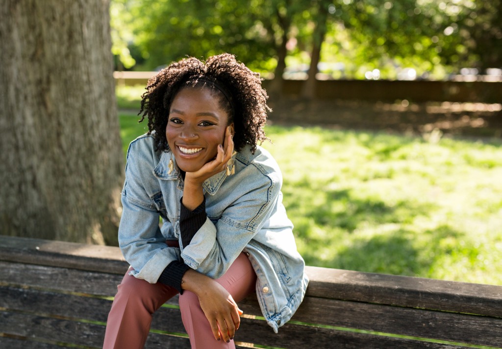 Alexa Patrick sits on a low stone wall in front of a tree in a park. She leans forward and smiles, resting her chin in her right hand. Alexa wears a jean jacket cuffed at the forearms with a black long-sleeved shirt underneath and dusky pink slacks. 