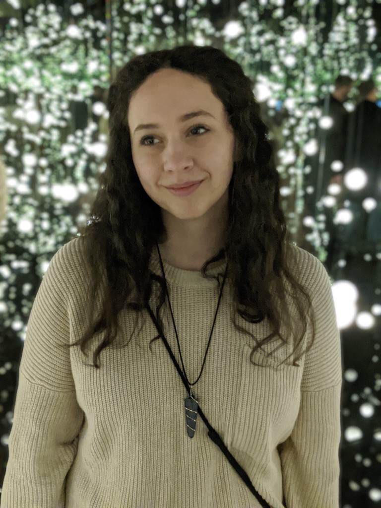 Alexandria, a white person with brown eyes and long, dark brown curly hair worn half pulled back, faces forward and looks to the left with a small smile. She wears an oatmeal-colored sweater with a pocket on the right side with a lapis lazuli pendant necklace. The black strap of their bag is worn diagonally across their torso from their left shoulder to their right hip. In the background, there are many white twinkling orb lights against a black backdrop.