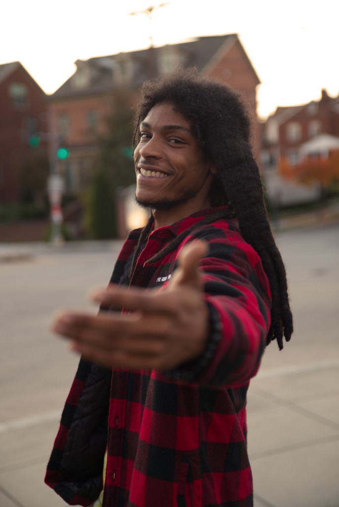 The camera faces the left side of Ayinde, who is looking towards the camera and smiling with his front arm reaching towards the lens in front of a residential intersection with two traffic lights on green and brick homes in the background. Ayinde is wearing a red and black checkered flannel with a chest pocket that reads “The World is Mind”, and a beaded black bracelet.  Ayinde’s skin is brown and his hair is long and dreadlocked in a freeform fashion, reaching below his front shoulder blade.