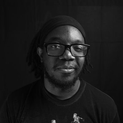 Brandon is shown in a black and white photo. His locs are tied toward the back of his head with a black headband. He is wearing glasses and dark colored shirt.