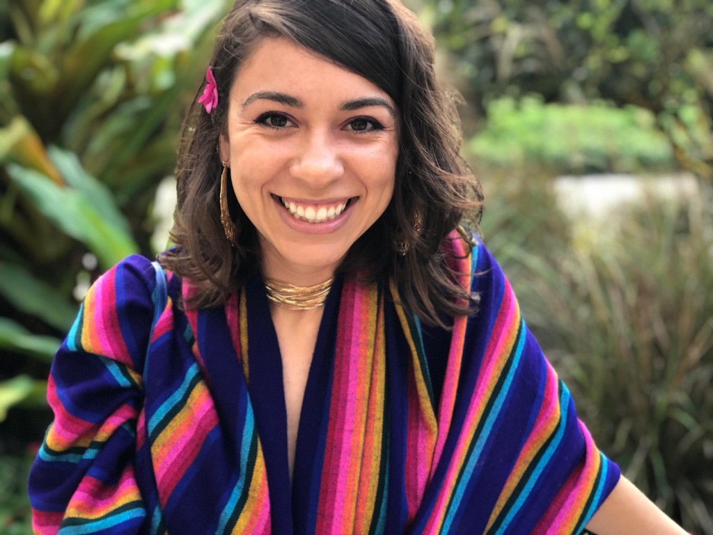 Chelsea appears outdoors smiling at the camera with tropical greenery in the background. She is white with dark brown eyes and dark brown, wavy hair that falls just below her shoulders. She wears a brightly colored, striped rebozo, gold hoop earrings, a gold necklace, and a pink flower in her hair.