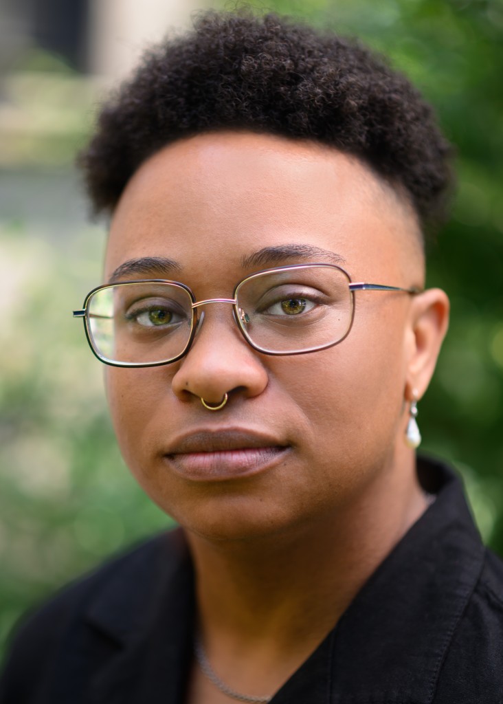 A lighter-skinned Black trans masculine person is photographed from the shoulders up, wearing a black lapel shirt with one dangly pearl earring, a gold septum ring, and rectangular glasses. He has a high top fade. The background is a blur of green bushes.