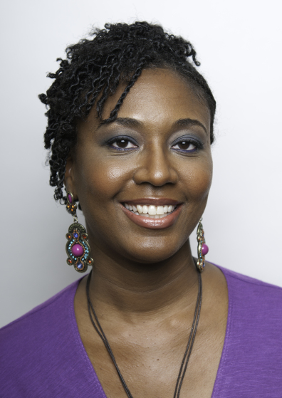 A close-up of Camisha Jones smiling. She has brown skin and her hair is styled in twists. She wears glasses, brightly colored earrings, and a necklace with a v-neck purple dress.