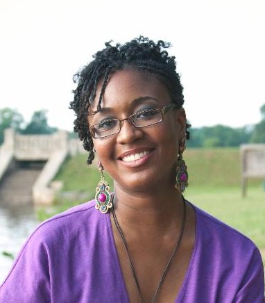 Camisha Jones smiles while sitting outside in a park. There is a body of water to her right and behind her there is grass, trees, and a bridge in the distance. She has brown skin and her hair is styled in two-strand twists. She wears glasses, dangling earrings decorated with jewel-toned stones, a necklace, and a v-neck purple dress.