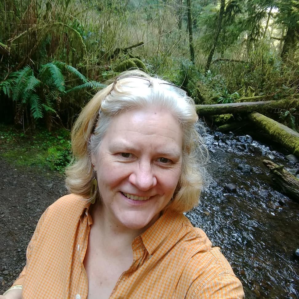 Sarah Browning appears outdoors while hiking. She wears an orange blouse and smiles toward the camera.