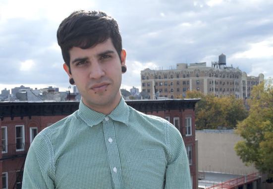 Image of Christopher Soto with fairly short dark hair, wearing a green stripped shirt buttoned all the way to the top, looking seriously towards the camera, and standing in front of a cityscape in the daytime.