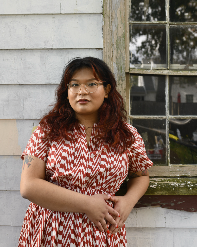 The poet Chrysanthemum stands outdoors and faces the camera with a wall and window behind her. She has golden skin and chest-length dark hair, and wears translucent glasses with a red-and-white patterned dress.