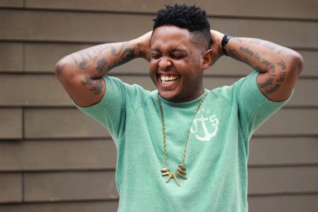 Danez Smith appears wearing a mint green t-shirt and a gold necklace. They hold both hands behind their head and laugh with their eyes closed.