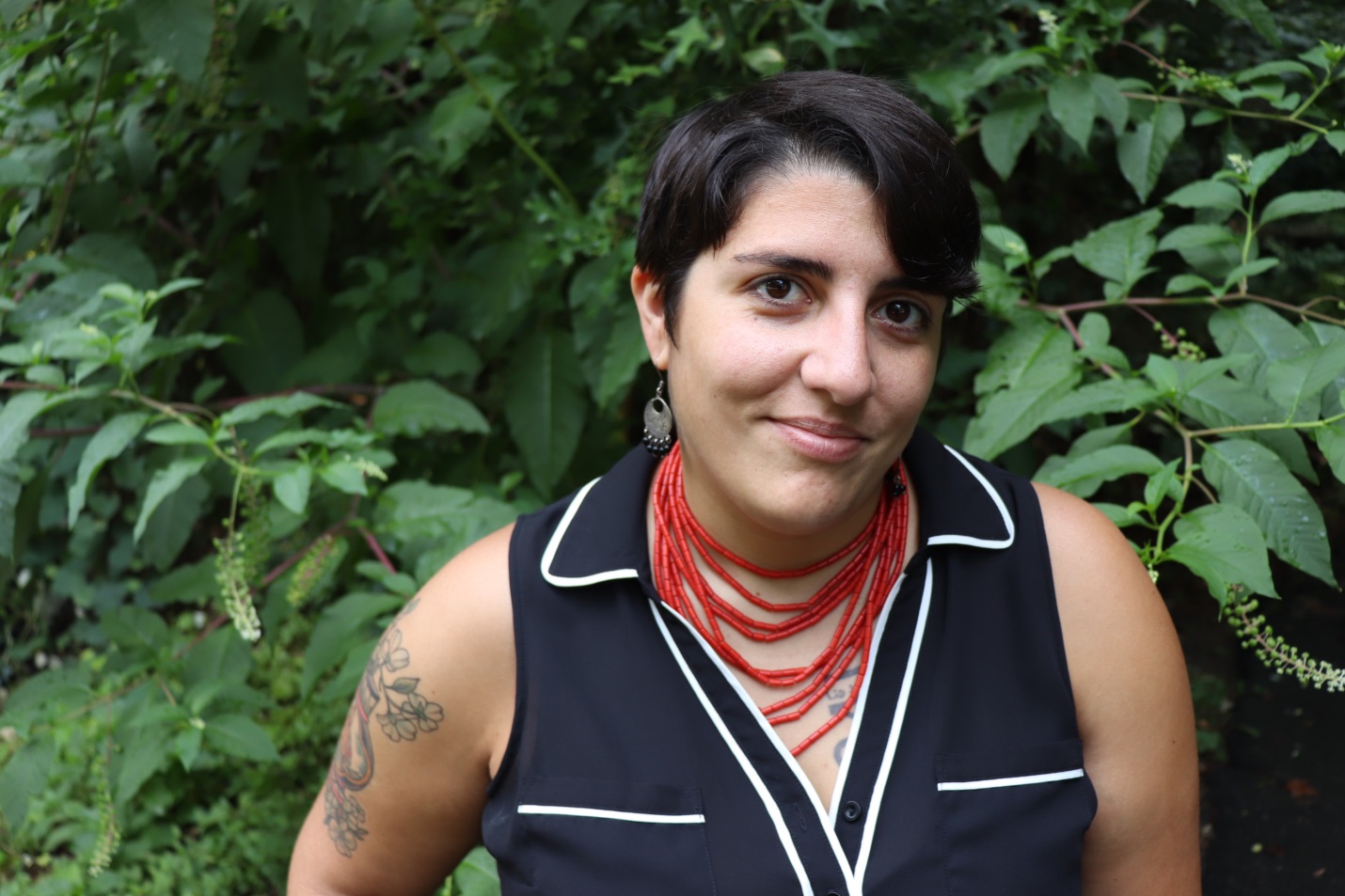 Danielle Badra, a queer Arab-American woman with dark short hair and multiple tattoos, in a black and white button up tank top with a red coral necklace and silver and black dangly earrings, stands in front of plush greenery. 