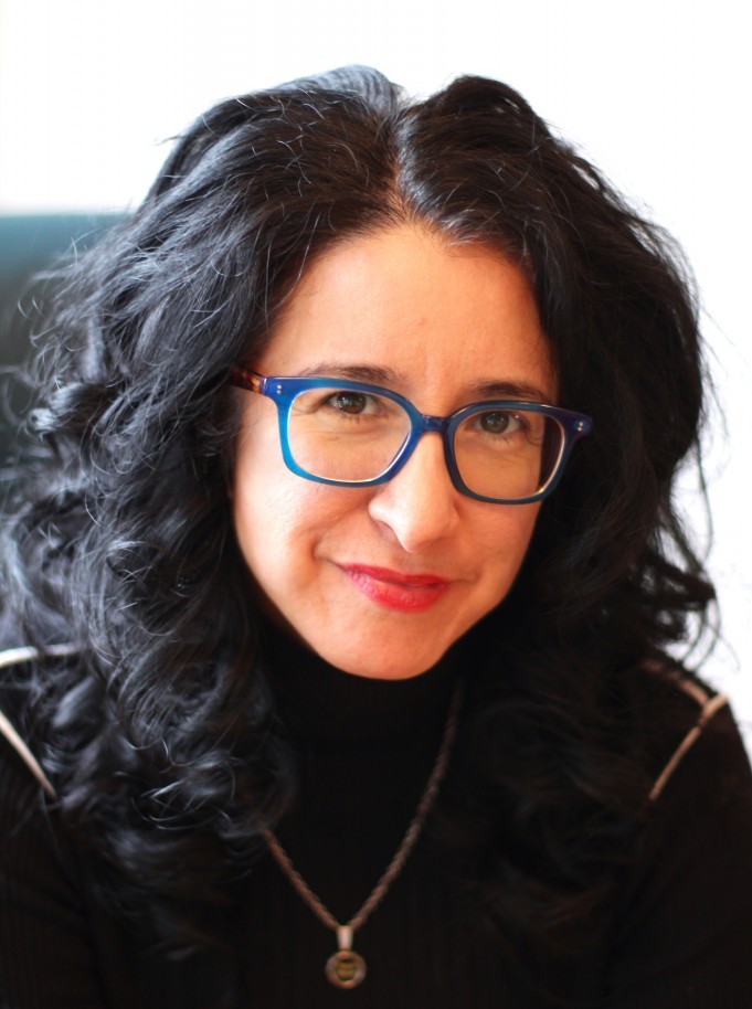 Close-up of Deborah Paredez, a woman with black wavy shoulder-length hair and blue-framed glasses wearing a black turtleneck and silver pendant necklace, seated in blue chair