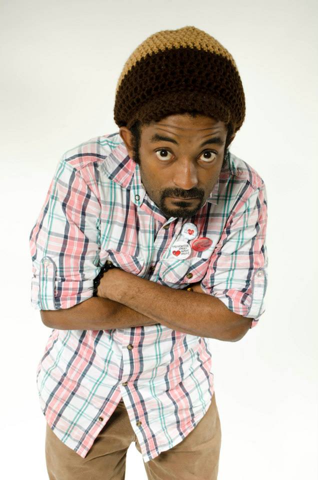 Dwayne Lawson Brown appears against a white background wearing a plaid short-sleeved button-up shirt and a brown and yellow crochet hat. He crosses his arms, leans over, and looks toward the viewer.