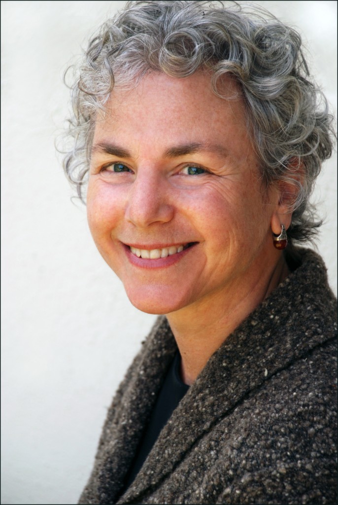 Image of Ellen Bass against a white background. Ellen has short, curly salt and pepper hair and green eyes. She wears a black shirt, grey wool jacket and silver earrings with a brown stone on the bottom. She is turned to the viewer’s left but smiling directly towards the camera.