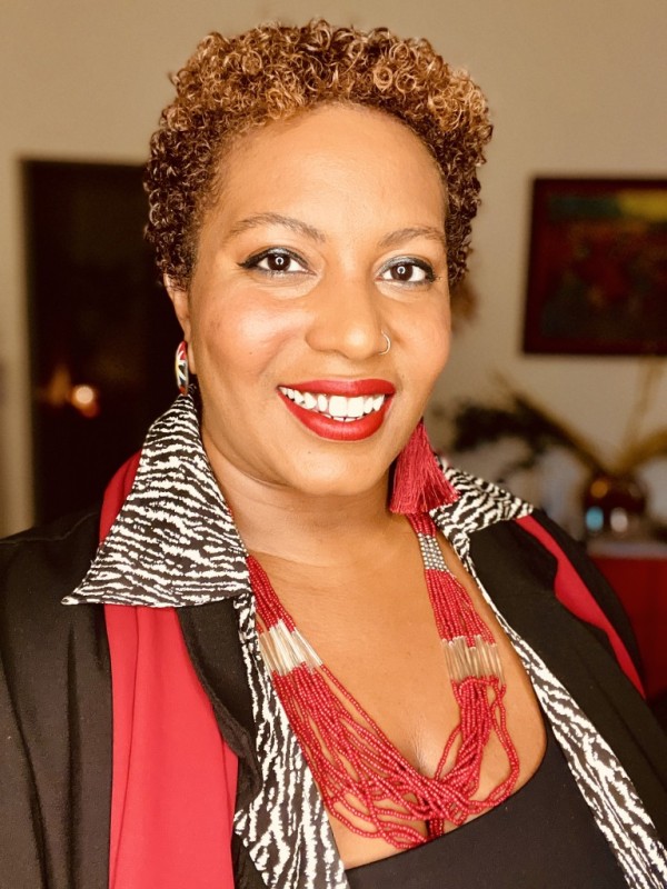 A person with short, wavy brown hair smiles into the camera. They are wearing a zebra-striped top, a red scarf, and a beaded red necklace. In the background is a painting of sharecroppers and a small ancestral altar with a burning candle.