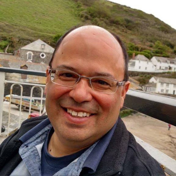 Photo of a Latino man with silver glasses and wearing a light blue shirt smiling into the camera with a grassy hill and homes behind him