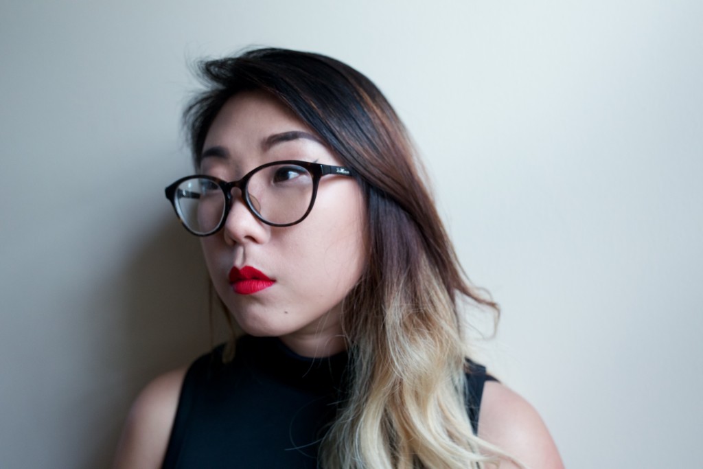 Franny Choi looks off to the right of the camera. Franny has long hair that is dark brown at the top and becomes lighter, eventually blonde at the bottom. Behind her is a white backdrop. She looks off into the distance. She wears glasses and a black tank top, and bright red lipstick. 