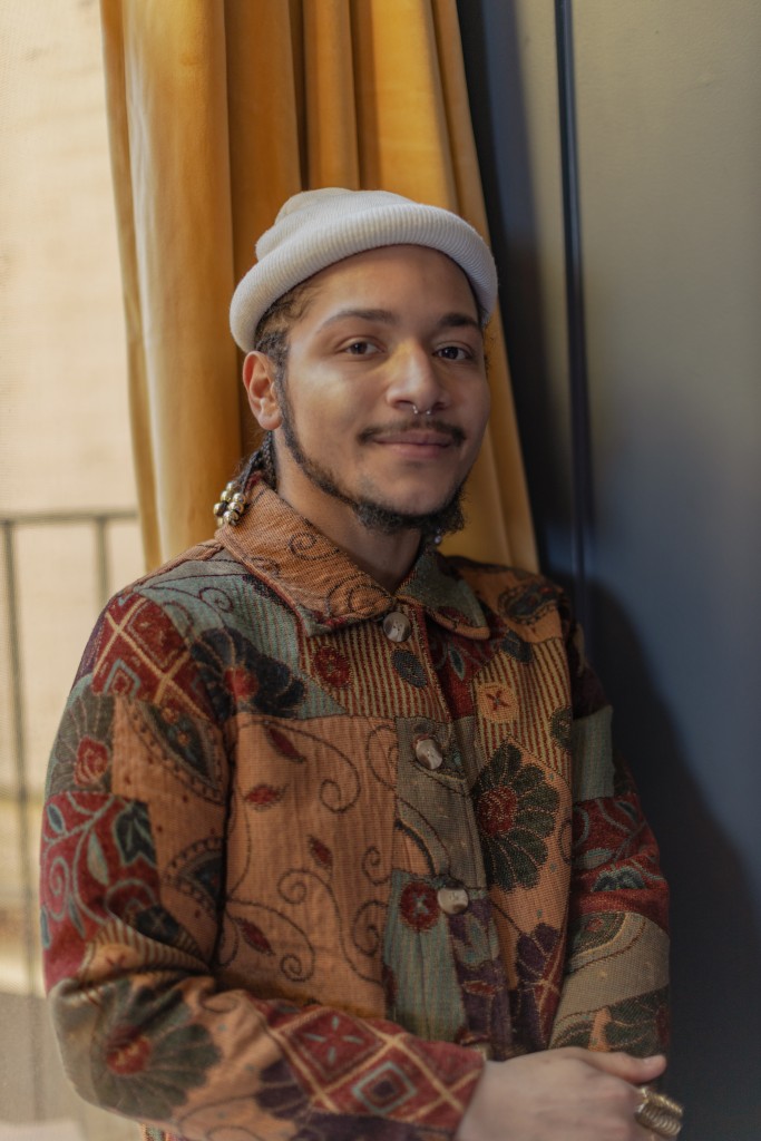 Gabriel Ramirez sits in front of a window partially covered with dark yellow curtains and smiles. He wears a white beanie hat, a patterned, collared, earth-tone button-up jacket, a septum piercing, and a gold statement ring.