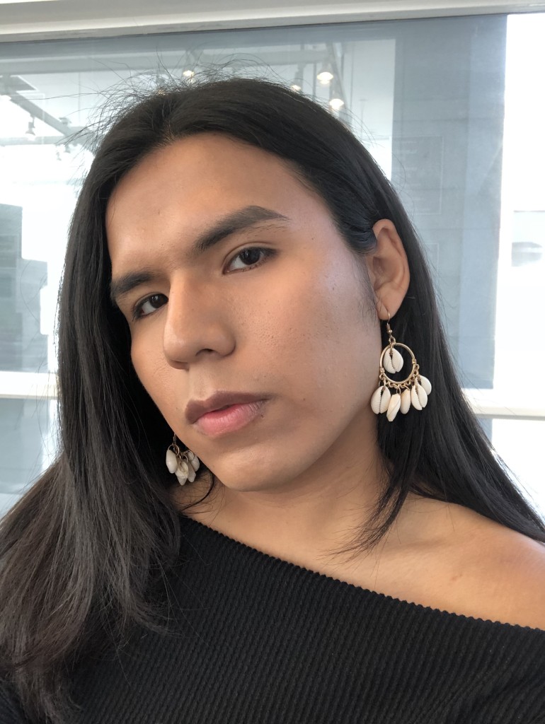 Jimena Lucero, a queer brown Latinx woman with her hair parted down the middle; she is wearing large dangly earrings, a black off the shoulder top, and has her face slightly turned away from the camera.