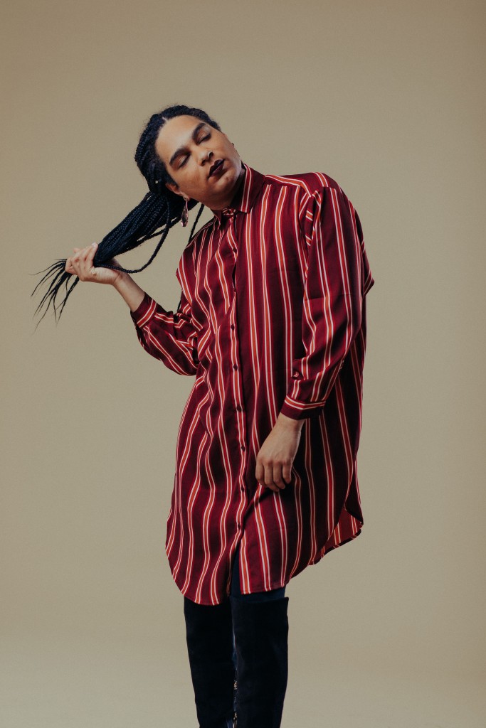 Justice Ameer in a red-striped shirt dress holding her braids with her eyes closed standing against a tan backdrop.