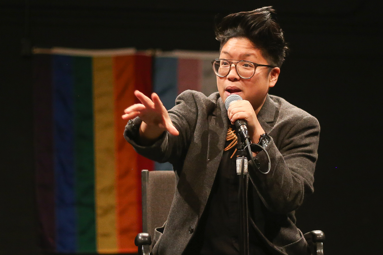 A light brown, round, queer performs at a microphone with transgender and rainbow flags in the background. They wear a gray blazer and glasses.