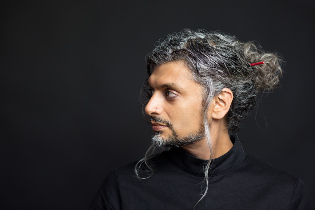 Image of Kazim Ali against a black background. Kazim wears a black turtleneck and looks slightly towards his right shoulder. He has a moustache, and a short goatee with side burns. His long, wavy salt and pepper hair is pulled back into a tousled bun held together with a red hair stick. His expression is neutral. 