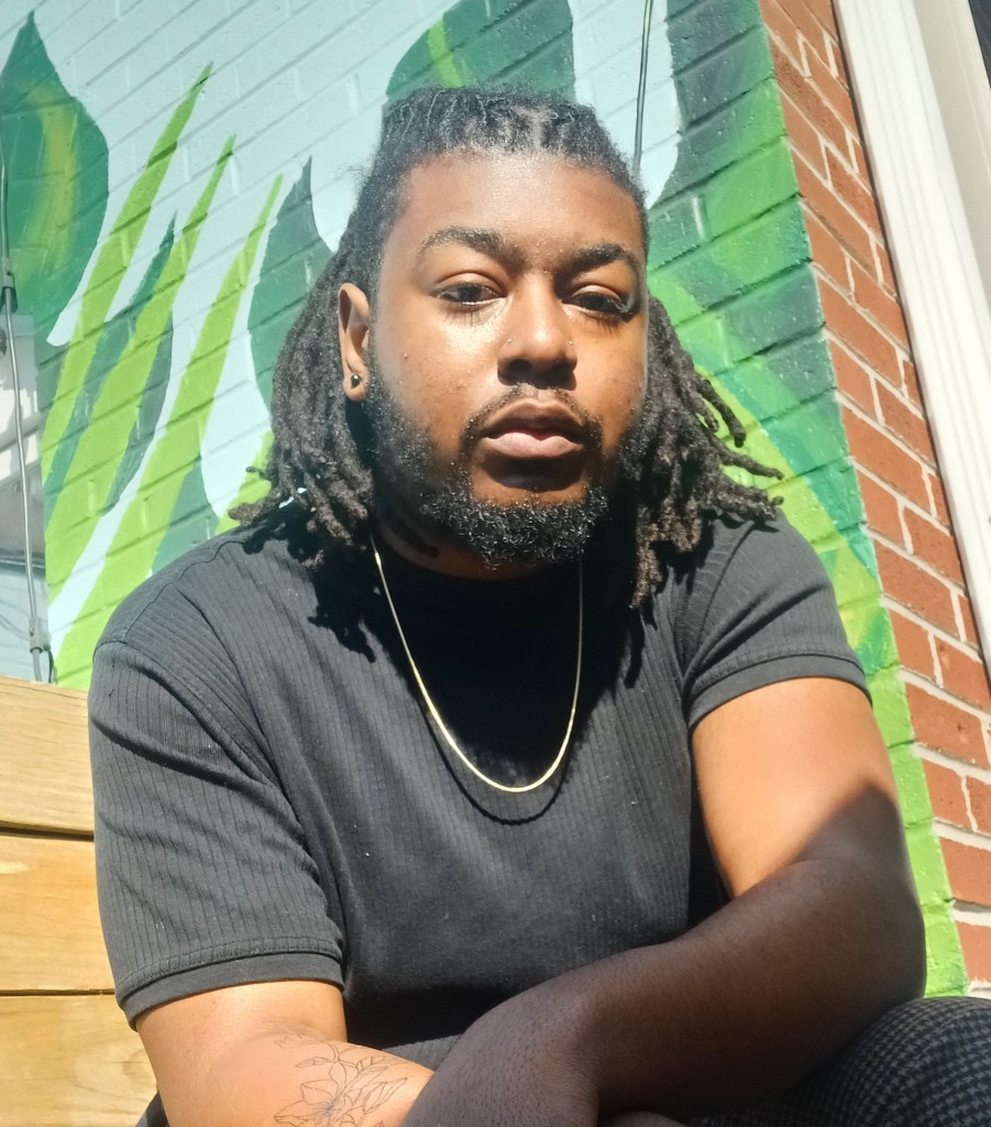 Kenny, a black man with long black locs that are braided back on his head, sits in front of a green mural on a bright day. He is wearing a short sleeve black shirt with a gold chain. He has a small tattoo of a magnolia on his right forearm.