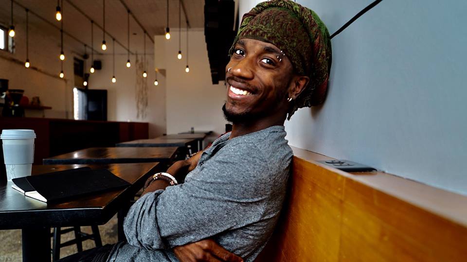 A man is seated in a coffee shop, sitting sideways to the viewer. He has his arms crossed lightly across his body, and his face is turned toward the viewer. He has brown skin and dark eyes and black goatee. He's wearing two piercings in one eyebrow and one in his earlobe. He's smiling widely at the viewer, and is relaxed against the back of the bench he's sitting on. He's wearing a light blue, long-sleeved Henley shirt, and a green turban with an intricate brown design on it.