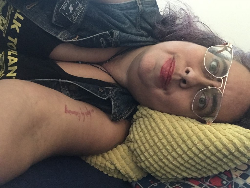 A 40ish mixed race Sri Lankan, Irish and Romani nonbinary  femme with curly brown silver and purple hair, lying on a couch looking at the viewer horizontally. They have rose gold aviator frames, thick eyebrows, red lipstick and sand colored skin, and are looking at the viewer with a kind of tired but hopefully crip wonder. They wear a blue denim vest with a pin that says Neurodivergent Universe above a pink and blue image of a ringed planet, and a black tank top with yellow lettering that read Talk To Plants, Not Cops is barely visible. They have a tattoo of the words 