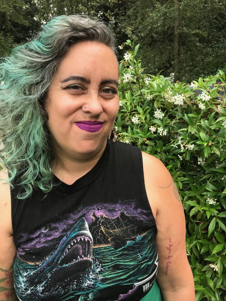 Leah Laksmi Piepzna-Samarasinha, a light brown skinned non-binary femme writer, looks at the viewer. She had long curly dark brown and green/teal hair with an undercut on her left side, magenta lipstick, and is wearing a black muscle t shirt with an imageof a shark rearing up out of the water on the front. She stands in a garden in South Seattle in late summer, with blooming jasmine behind her.