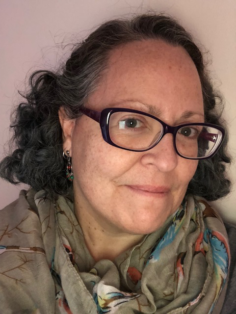  Portrait image of M. Soledad Caballero. She wears dark-framed glasses, a beige scarf with a floral pattern, and drop earrings with multicolored beads. She has dark hair with shocks of gray at her temples, and light brown eyes.