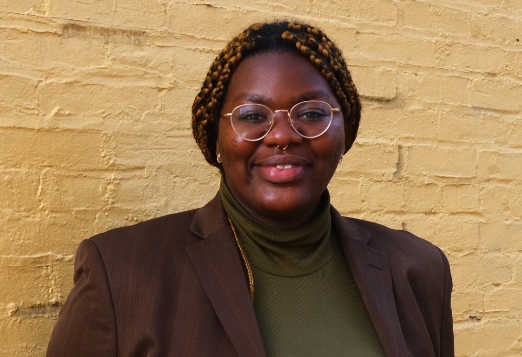 M Kamara stands in front of a beige painted brick wall. They wear a dark brown blazer with a green turtle neck, glasses, small gold earrings, and a septum piercing. They wear their hair in braids pulled back from their face.  