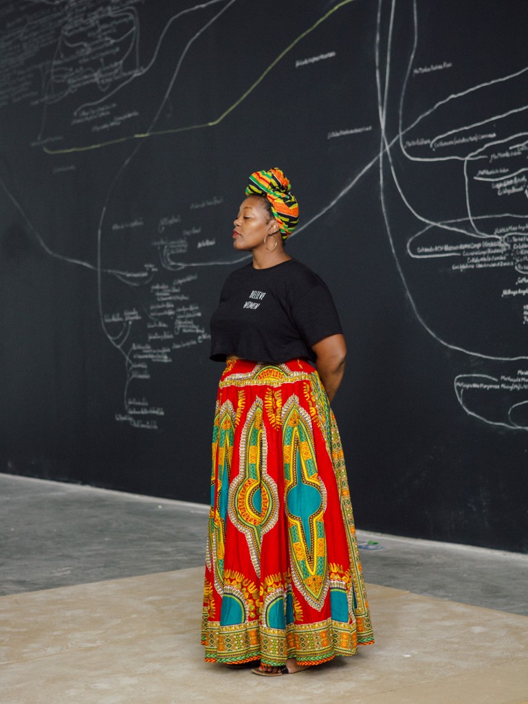 Mahogany L. Browne wearing a black shirt and colorful skirt standing in front of a chalkboard.