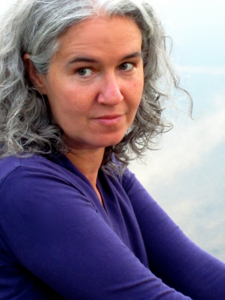 Photo of Melissa Tuckey wearing a purple long sleeve shirt. She is seated turned to the left and looks toward the camera over her right shoulder. Her gray wavy hair is slightly parted in the middle tucked behind her right ear. Her expression is neutral.