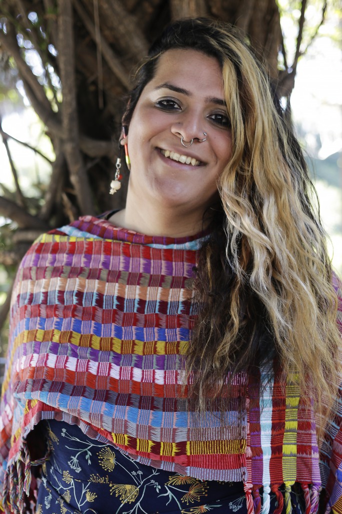 Moncho Alvarado, a queer trans woman of color with bold blond and dark brown hair, is smiling widely. She wears a multi-colored shawl with a colored pencil and a dangling earring in their right ear as well as two circular nose piercings. Her hair is worn swept over her left shoulder. She is outside with branches in the background.