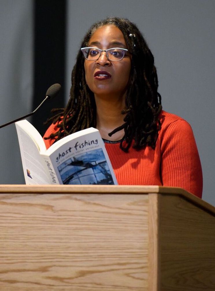 Camille Dungy reads from the Ghost Fishing anthology standing at a podium. She is a black woman with long locked hair, glasses, and an orange shirt.