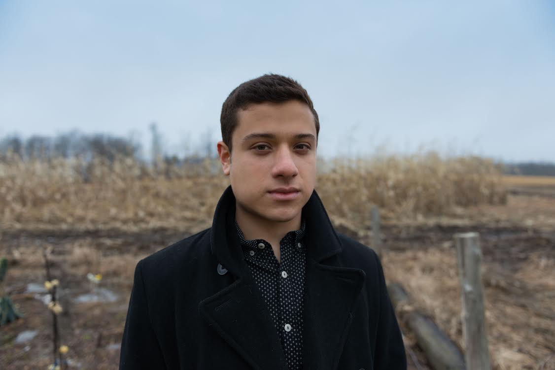 Image of Jonathan Mendoza standing near a winter corn field. The sky is blue and overcast. He looks directly at the camera, wears a heavy brown overcoat, and has short dark brown hair and brown eyes.