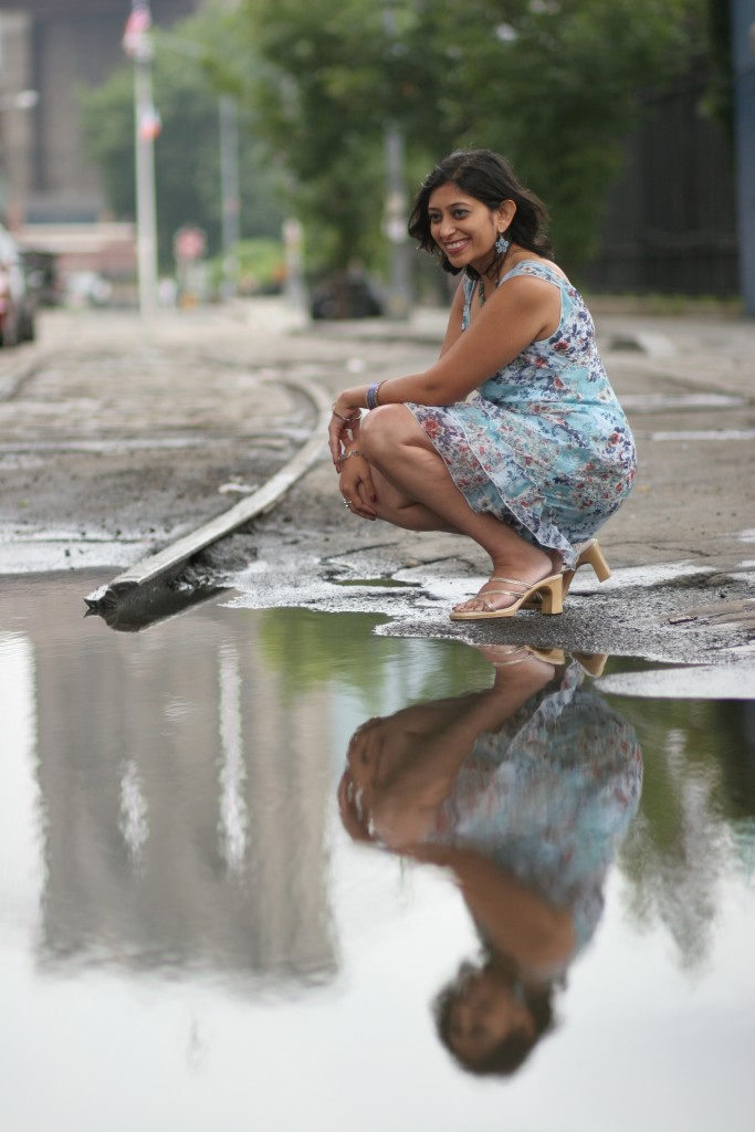 Photo of Purvi Shah, an Indian woman in a blue and purple dress kneels down next to a puddle smiling widely
