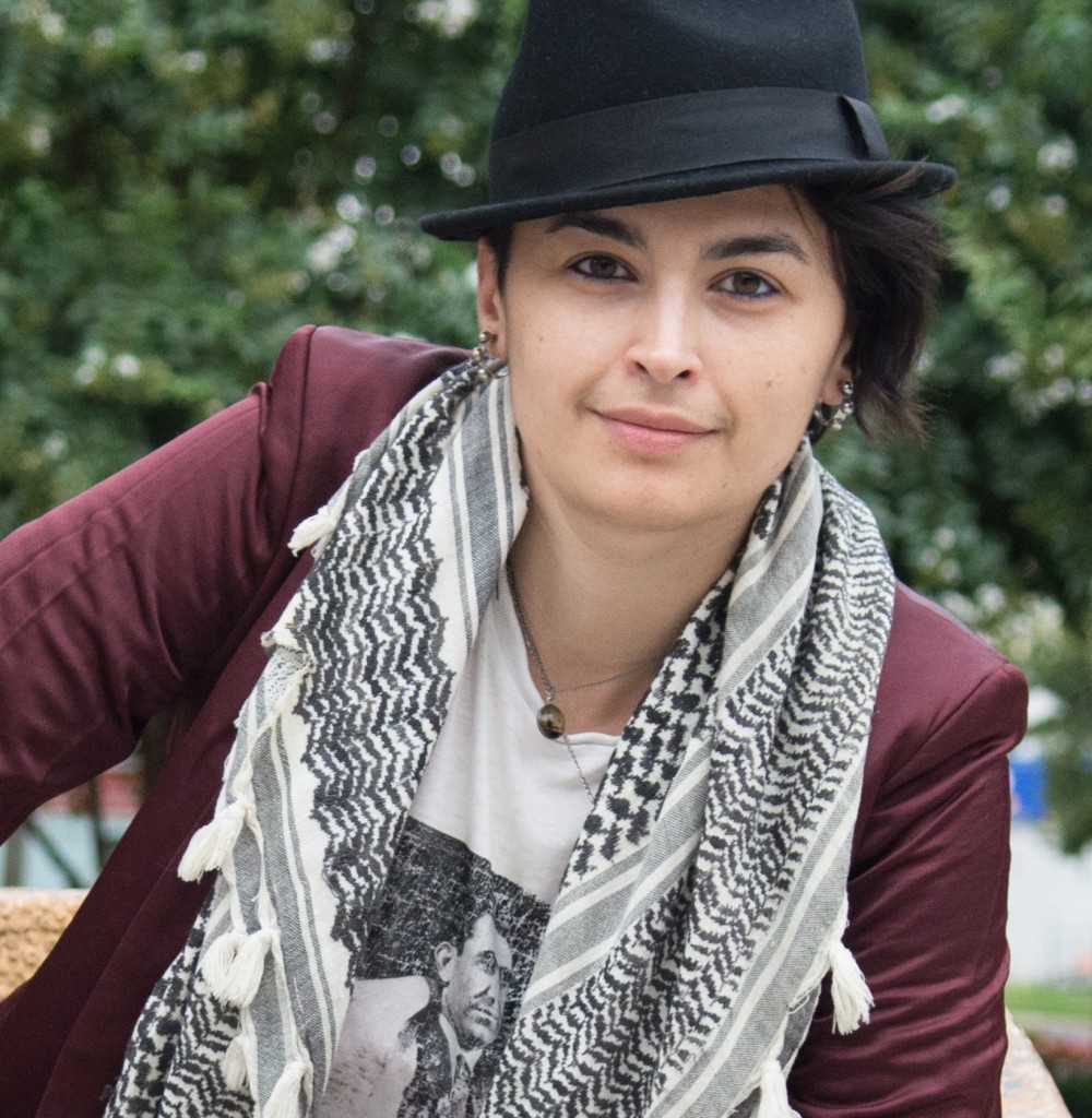 Poet and organizer Rasha Abdulhadi looks directly at the camera. They wear a black fedora and a wine colored blazer and a black and white scarf.