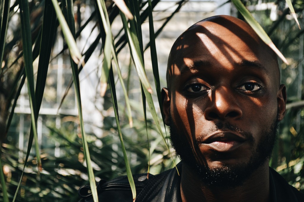 Image of Rasheed Copeland standing amongst thin palm leaves. He has a neutral expression and the shadow of the leaves appear across his face.