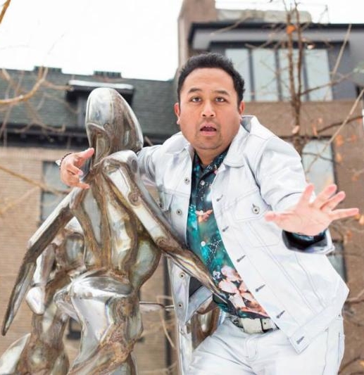 Photo of Regie Cabico in white jacket with arm around a sculpture