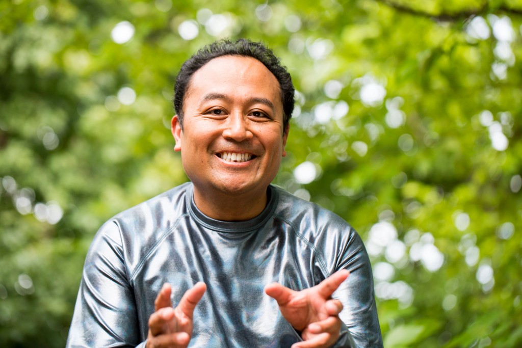 Regie Cabico appears outdoors with greenery in the background. He wears a silver shirt and smiles as he gestures with both hands toward the viewer.