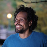 Image of Ross Gay. He wears a blue shirt and smiles. Blurred in the background are trees and a light post.