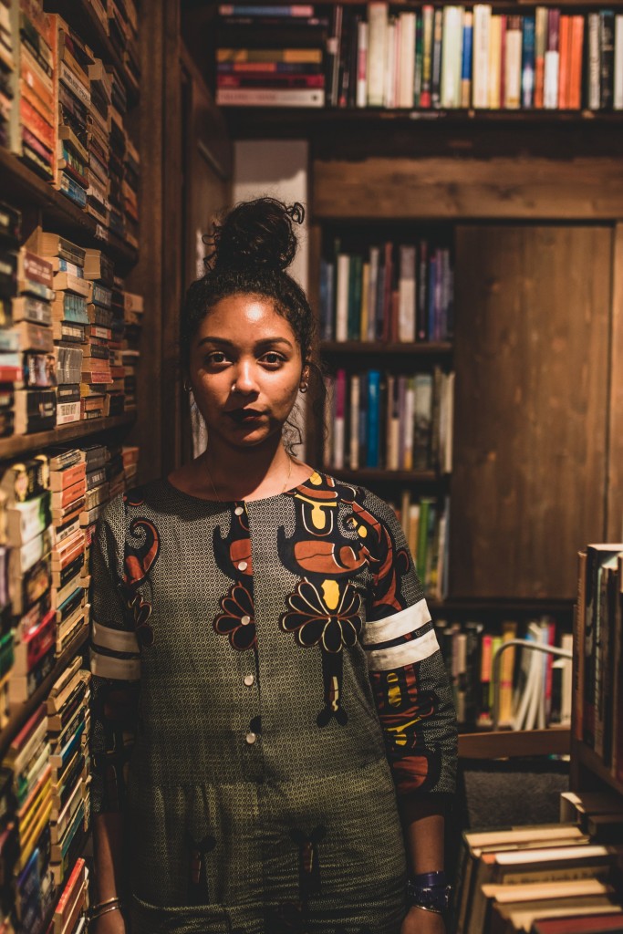 Safia Elhillo front facing looking into camera next to a shelf full of books.