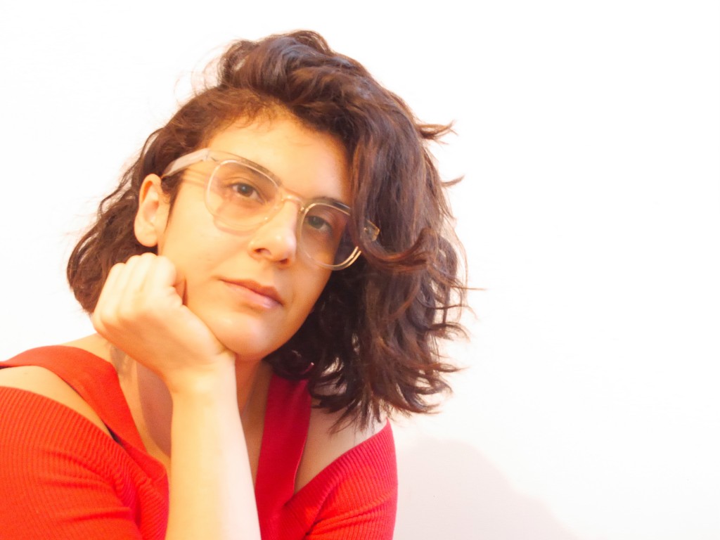 Shabnam Piryaei leans her chin on her hand and looks towards the camera. She has medium length wavy brown hair worn parted and flipped to the side. She has brown eyes and is wearing glasses with clear frames. She wears a cherry red top. 